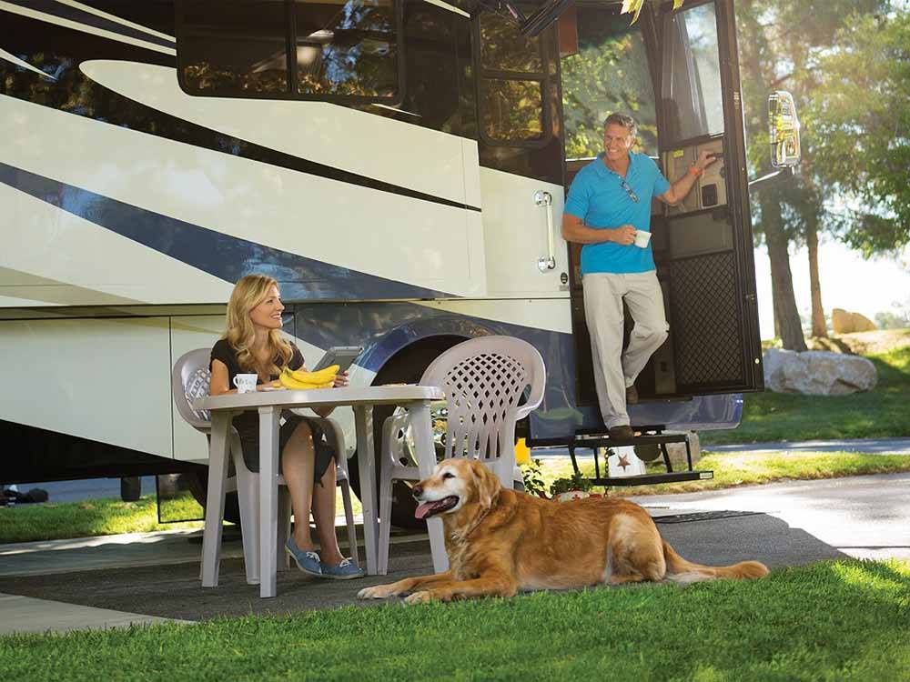 A dog sitting in front of a woman with a man holding a cup exiting a Class A RV at LAKESIDE CASINO & RV PARK