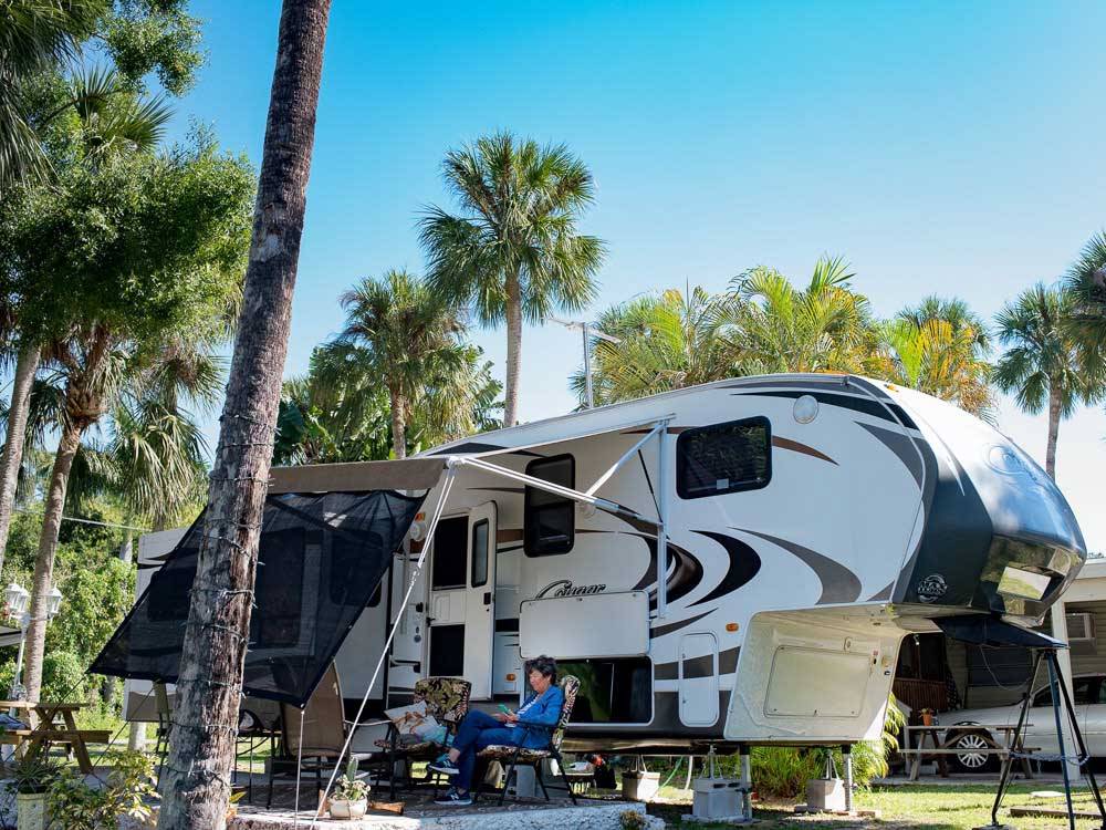 Two campers relax outside of a fifth-wheel amid palm trees at BLUEWAY RV VILLAGE