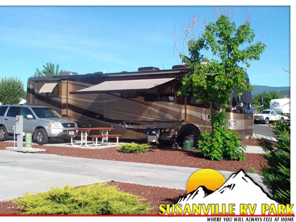 Large brown and tan RV and SUV parked at a paved site at SUSANVILLE RV PARK