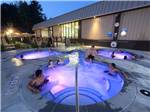 Outdoor hot tub lit in purple hues at night with several people relaxing in it at LAKE GEORGE RV PARK - thumbnail