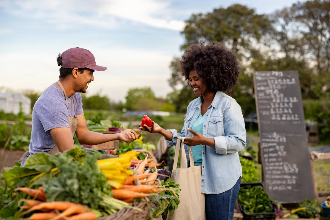 Vendor handing fresh produce to a smiling customer at an outdoor farmers market.