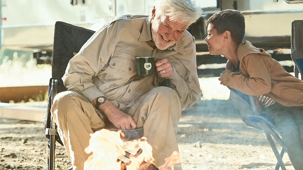 An older man and a child laughing together by a campfire in front of an RV