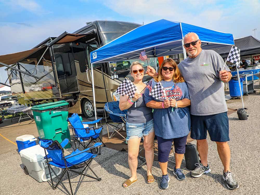 Three people standing in front of their RV with racing flags at WORLD WIDE TECHNOLOGY RECEWAY CAMPGROUND