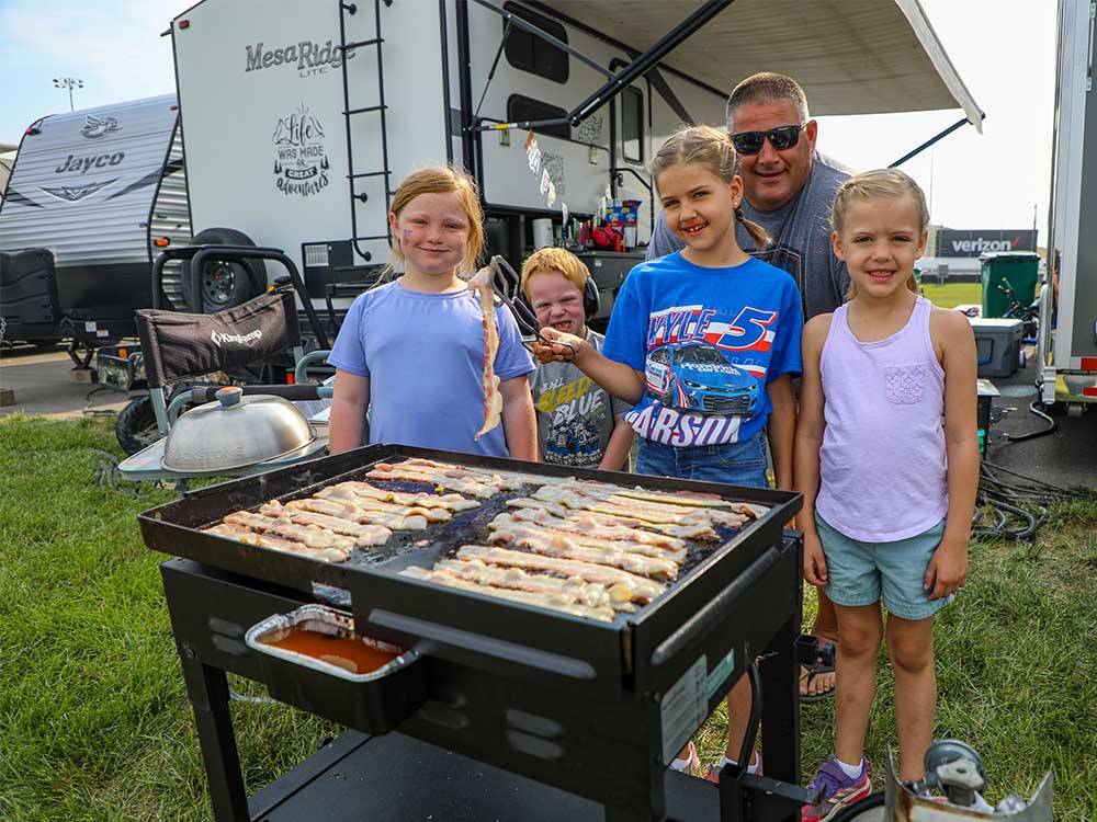 A dad and his children cooking bacon on the grill at WORLD WIDE TECHNOLOGY RECEWAY CAMPGROUND