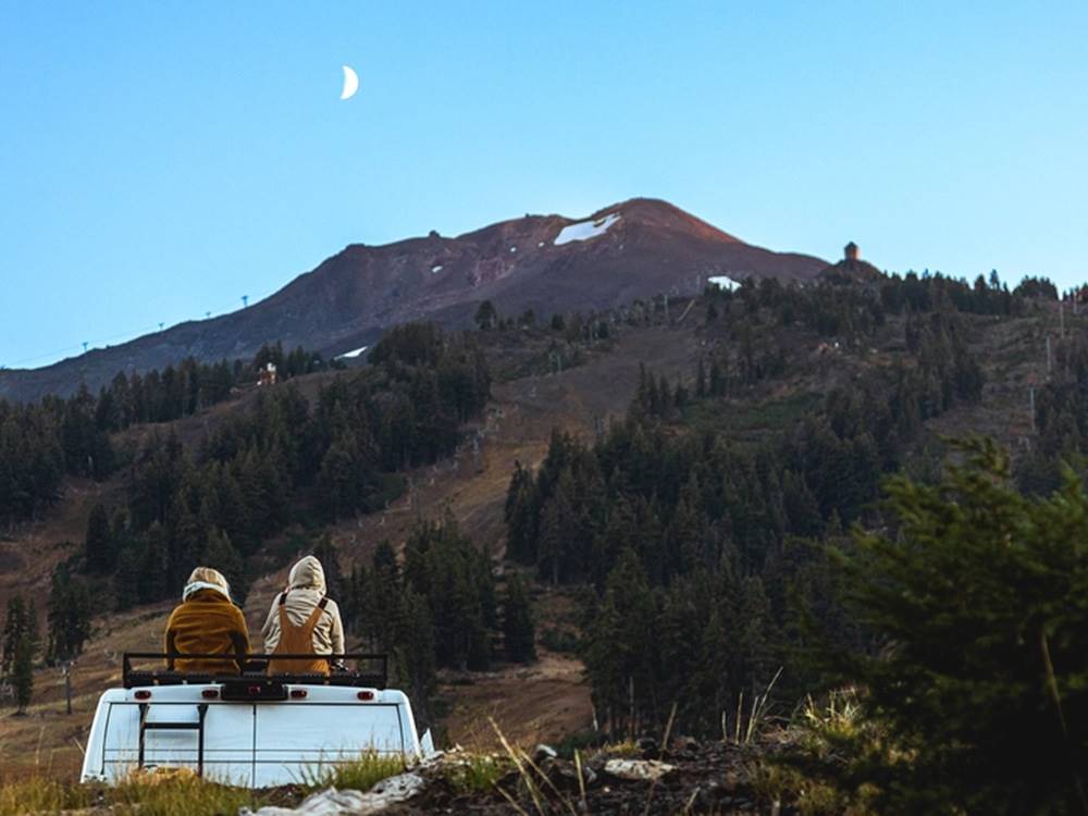 Two people sitting on RV with mountain and moon
