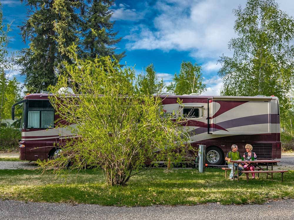Motorhome in campsite with couple at picnic table at RIVER'S EDGE RV PARK & CAMPGROUND