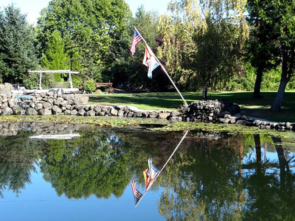 Trees, lawn with flags flying over pond at ON THE RIVER GOLF & RV RESORT