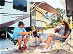 Family sitting outside their RV on a sunny day at an RV site - thumbnail