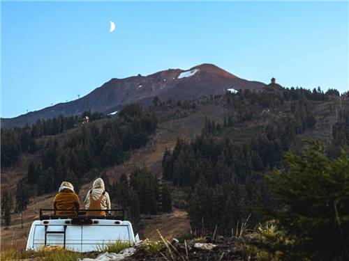 Two people sitting on RV with mountain and moon